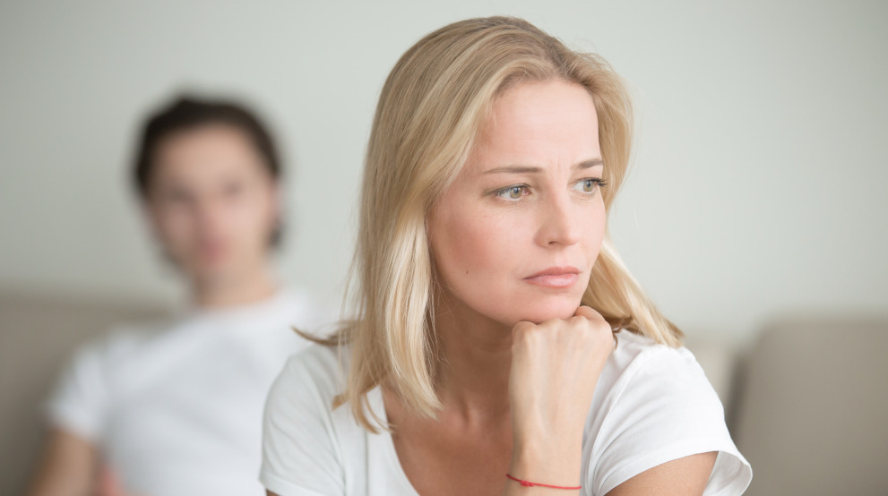 Frau nachdenklich und verzweifelt, im Hintergrund sitzt Ihr Mann - eine Affäre ist ein tiefer Einschnitt in die Beziehung